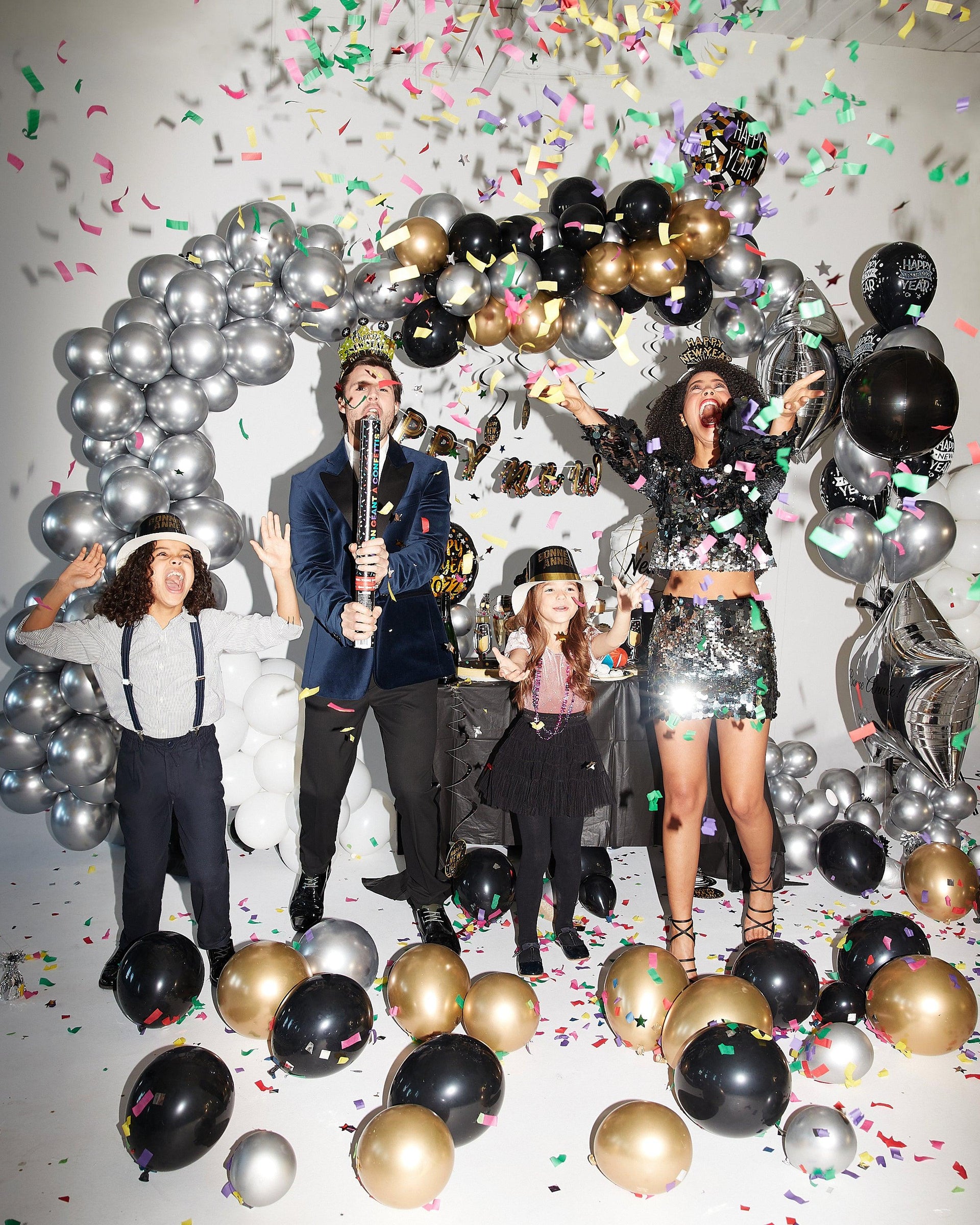 A woman, a man, a little girl and a little boy are celebrating New Year's Eve together. They have a lot of fun surrounded by many balloons, decorations and confetti. The colour theme is silver, gold and black and it is very sparkly.